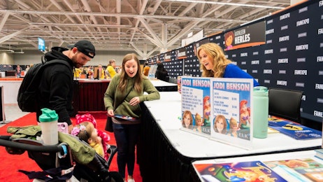 The image captures a heartwarming moment at FAN EXPO where a young family is interacting with Jodi Benson at her signing booth. On the left, a man in a black hoodie and cap tends to a toddler in a stroller, who is dressed in a cute, yellow outfit. Next to him, a woman in a green sweater looks surprised and delighted as she talks to the celebrity, Jodi Benson, known for voicing a popular animated character. Jodi Benson, behind a booth decorated with colorful signage of her name and character images, is smiling broadly at the toddler, adding to the joyful atmosphere. This scene is set against a backdrop of the expo's vibrant environment.