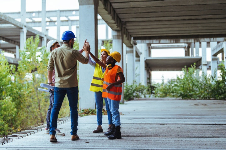 A joyful group of diverse construction workers exchange high-fives on a building project site, wearing hard hats and safety vests, with blueprints in hand.