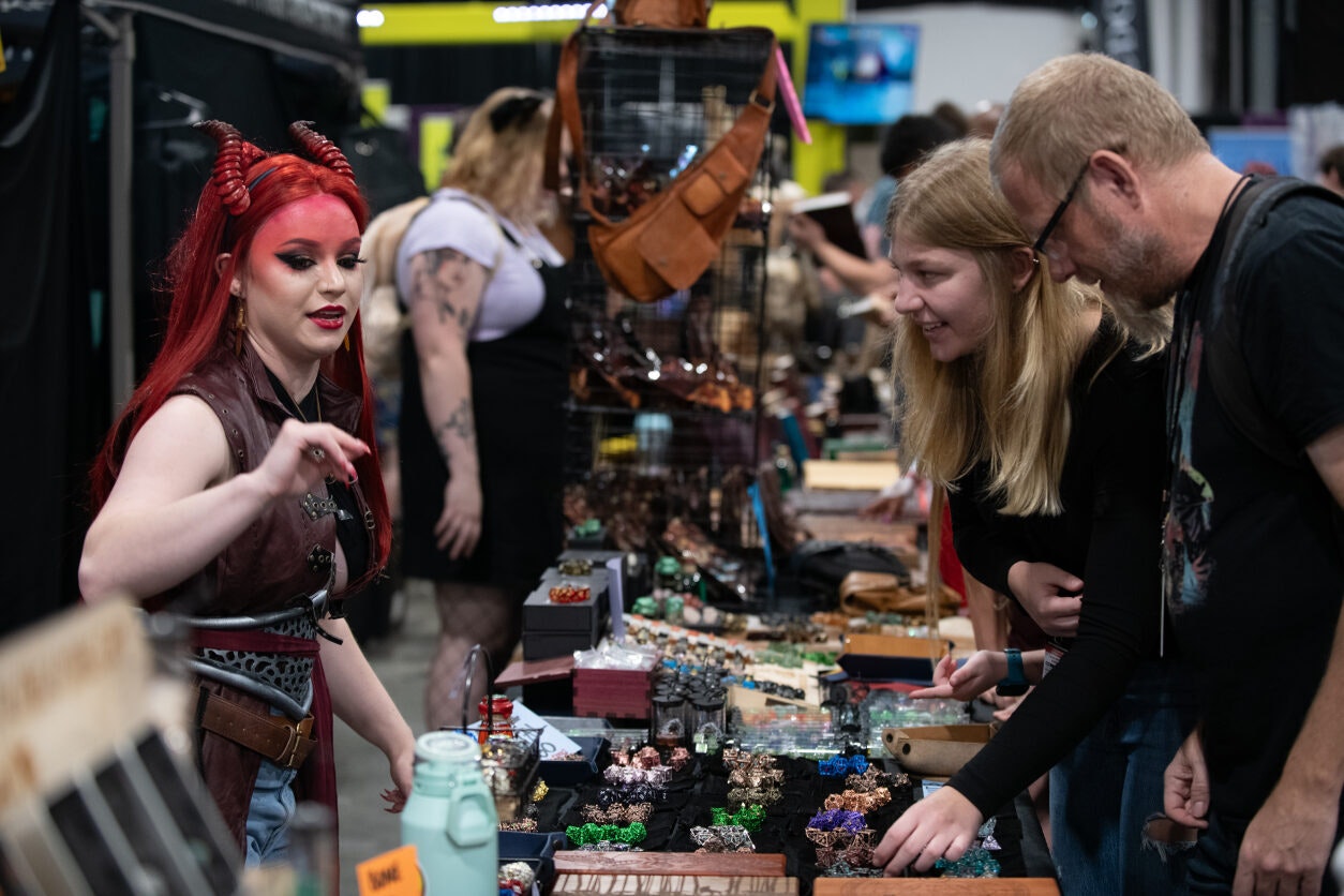 Dressed as a teifling, the person behind the Misty Mountain Gaming booth helps a couple find a new set of dice