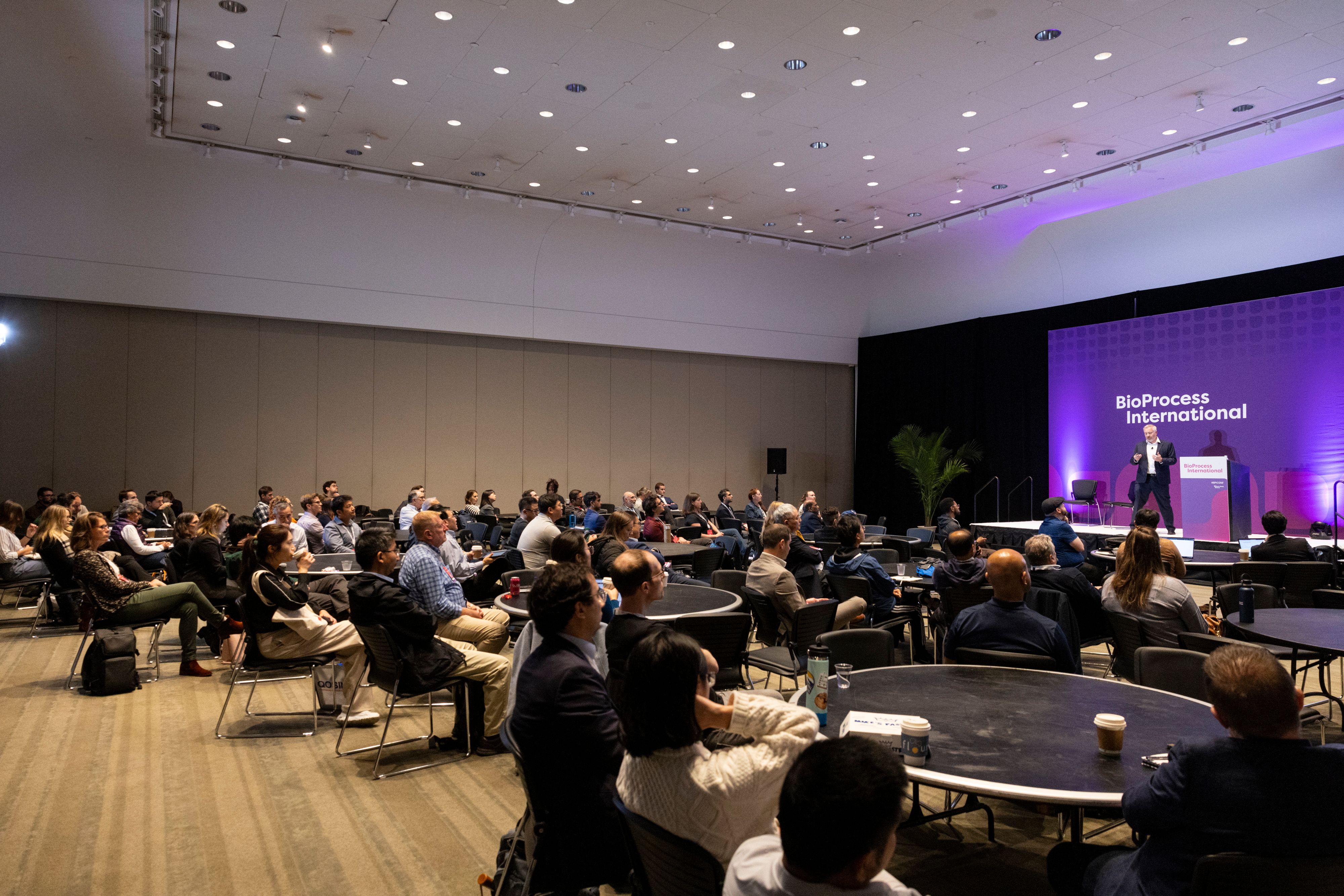 A speaker leads a talk in a room of bioprocessing professionals sat around round tables at BioProcess International (BPI).