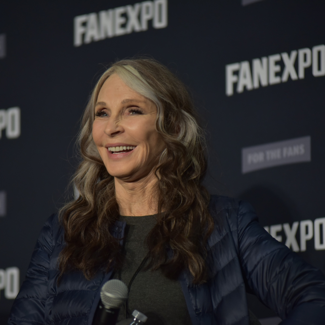 Gates McFadden smiling on a panel at FAN EXPO. She is wearing a gray shirt and a blue puffy coat.