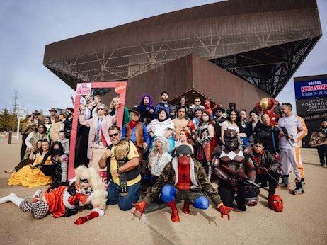 A diverse group of individuals dressed in various costumes gather outside a large convention center. The costumes represent characters from comics, movies, and other popular culture references. Notable outfits include superheroes, soldiers, a Barbie doll in its packaging, and fantasy characters. A banner in the background reads "Dallas FANFESTIVAL October 20-22," suggesting the group is attending a fan convention. The atmosphere is festive, with participants posing proudly in their outfits against the architectural backdrop of the convention center.