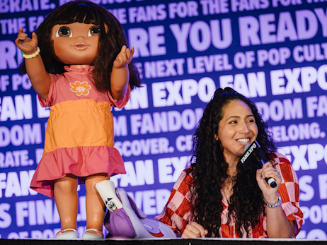 Kathleen Herles smiling, holding a microphone on stage, with a large, life-size doll of Dora The Explorer on her left.