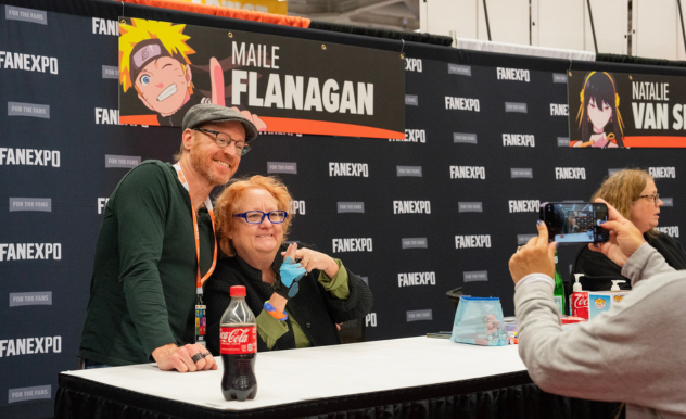 The image features a heartfelt moment at a FAN EXPO event, where Maile Flanagan and Chris Edgerly are posing for a photograph at a booth. Maile is warmly embracing Chris. She is smiling joyfully and dressed casually. Chris is leaning close to her with a delighted smile, wearing glasses and a green t-shirt. Another attendee captures this moment with a smartphone, adding a personal touch to the interaction. The booth's background is adorned with promotional materials, creating a vibrant and engaging atmosphere.