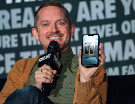The image features Elijah Wood at a FAN EXPO panel, smiling broadly as he holds a microphone in one hand and a smartphone displaying a photo in the other. He is dressed in a casual olive-green shirt under a mustard-colored cardigan. The photo on the phone shows two candles, seemingly from a movie or show, adding a layer of interaction or reference to his discussion. The background is filled with the FAN EXPO logo repeated across a dark backdrop, emphasizing the event's branding. This setting suggests he is engaging the audience, perhaps sharing a story, or discussing a topic related to the content shown on his phone.