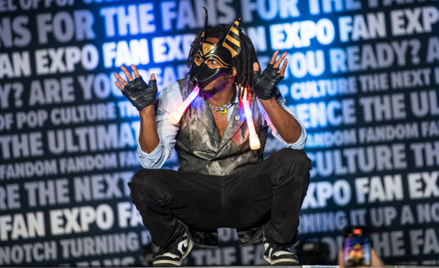 The image features an individual crouched on a stage in a dynamic pose, dressed in a costume with futuristic or tribal elements. The costume includes a vest, pants, and sneakers, paired with a mask adorned with stripes and glowing elements. Their hands are gloved, and they appear to be holding light sticks, adding to the performance aspect of the costume. The person's pose is energetic and engaging, suggesting they might be in the midst of a dance or act.