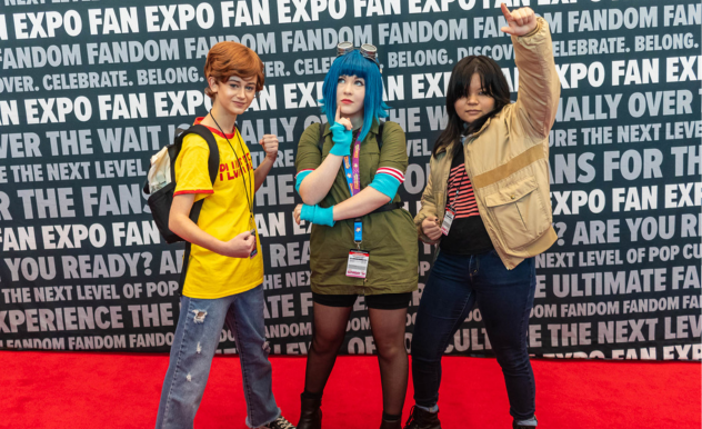 The image depicts three individuals striking poses on a red carpet against a backdrop with "FAN EXPO" branding, indicative of a fan convention. Each person is dressed as a character that might be found in a modern or urban setting, as opposed to a fantastical one. The individual on the left is wearing a yellow T-shirt with "PLUTO" printed on it, jeans, a backpack, and a wig with short brown hair, giving off a youthful, school-kid vibe. The person in the center has a blue wig, gloves, and a green dress, adopting a superhero-like stance with a fist raised. The one on the right is dressed in a casual jacket and striped T-shirt, with dark hair covering part of their face, embodying a cool, nonchalant attitude. Together, they reflect a variety of characters one might see in contemporary animated series or graphic novels.