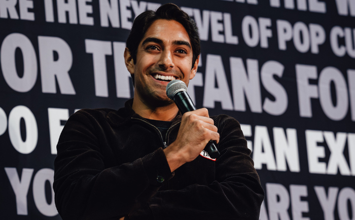 Eman Esfandi with dark hair and a bright smile speaks into a microphone. He's wearing a black zip-up jacket and has a warm and approachable demeanor. He's positioned in front of a backdrop with repeated fan expo text, suggesting he's participating in a panel or event.