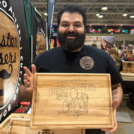 Owner of Histper Lasers smiles for the camera as he holds up one of their X-Men themed cutting coards. The light wood board features a Charles Xavier image, where his head is an egg, and the words "Professor Eggs-avier"