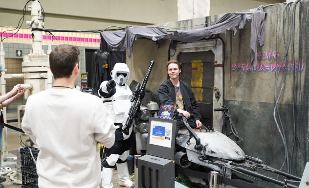 A fan takes a photo of their friend sitting on the speeder at the 501st booth. A trooper stands next to them pointing at the camera.