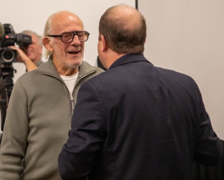 Christopher Lloyd smiles as Colorado Governor Jared Polis as they discuss the impact of pop culture on the Governor's life. Channel 9 films for the news in the back.