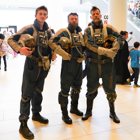 3 men, dressed as X-wing pilots, stand stoic as they stare at the camera, holding their helmets under their arms