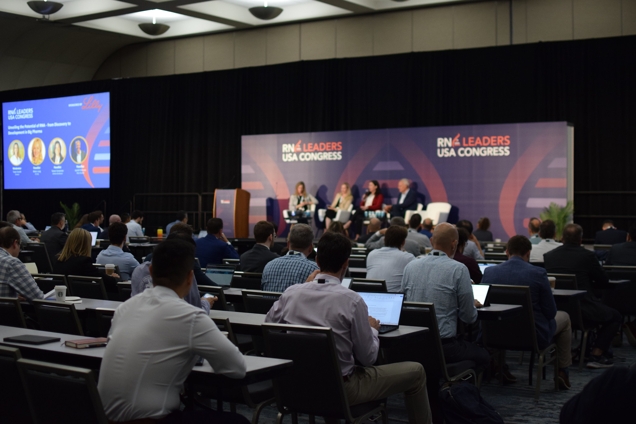 In a conference room, four panel speakers sit on stage whilst the audience watch, seated.