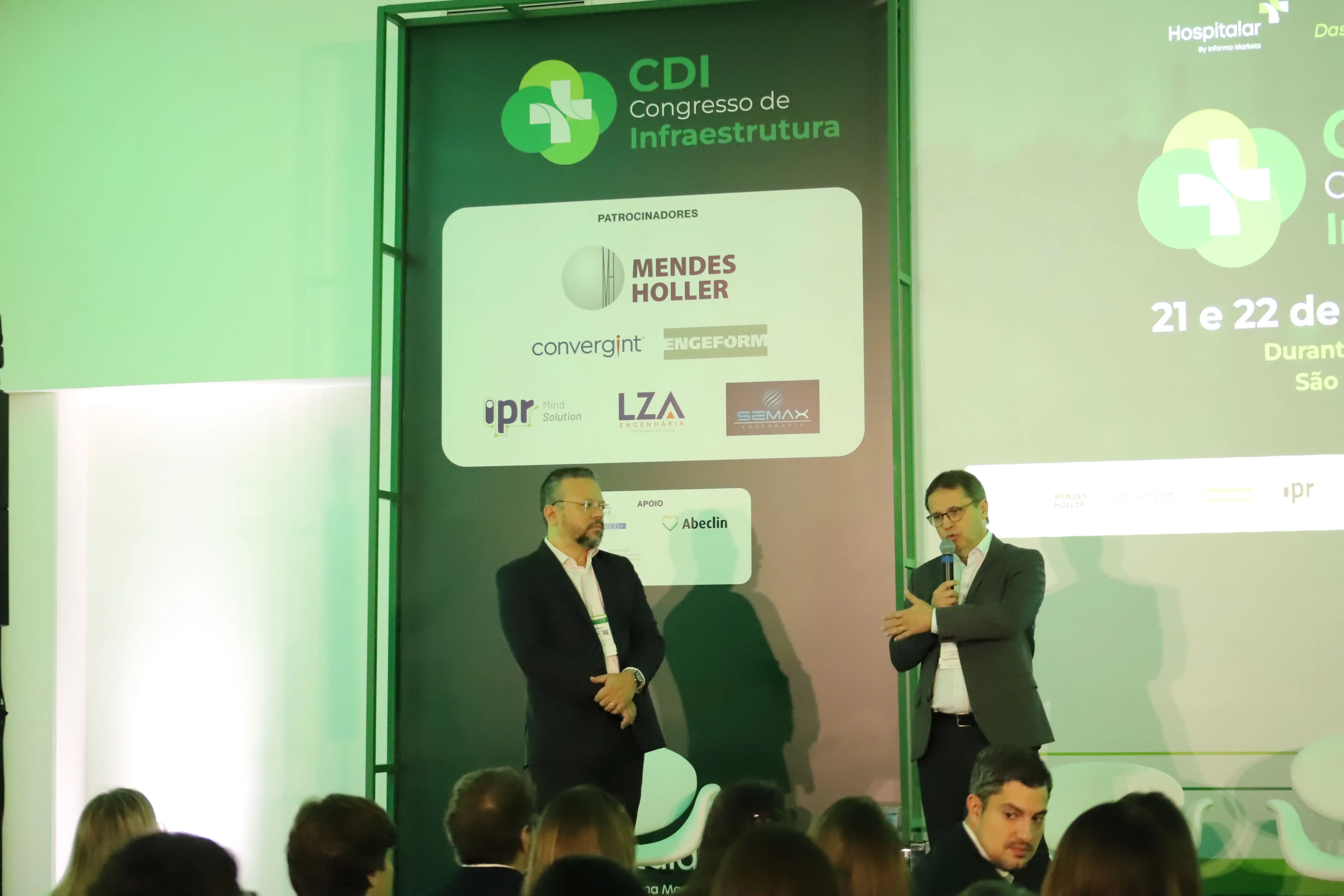 Vista frontal do palco do Congresso CDI. Um mediador e um palestrante estão em pé em um palco realizando uma entrevista. No telão ao fundo, o logotipo do 'CDI Congresso de Infraestrutura'. Atrás dos homens há um banner exibe as marcas patrocinadoras e o logo da Hospitalar.