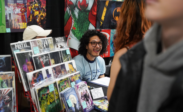 Isaac Goodhart greets fans at his booth in the Comic Book zone. He smiles as he chats with the fan facing him.