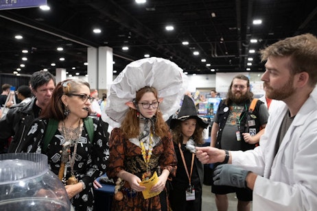 A family, dressed as gothic forest dwellers (witch, mushroom faerie, and another witch) listen as a person in a white lab coat shows them an unidentifiable object