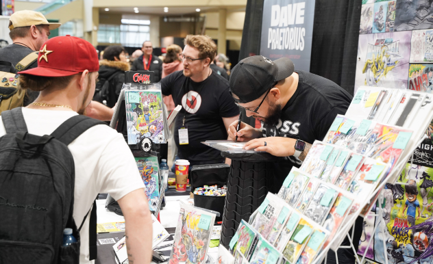 Comic artist signs one of his works for a fan waiting at his table in the expo hall. He is surrounded by comics for purchase. 