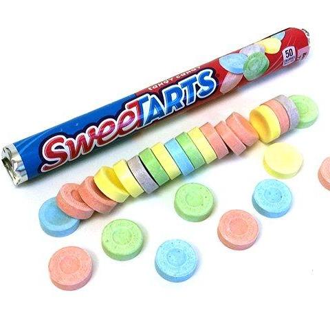 A tube of SweeTarts, pink and blue with writing in the reserve colours. In front of the tube is a row of the candies in red, green, blue, yellow, and purple.