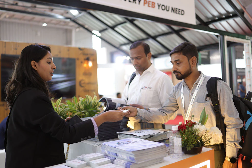 Exhibitor presenting products to several attendees at a table inside a booth during Roof India 2025
