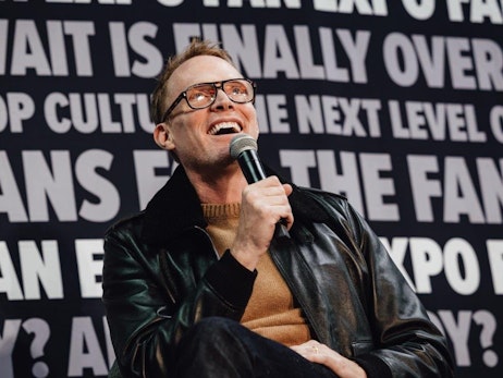Paul Bettany with glasses, wearing a brown sweater and black leather jacket, holds a microphone and laughs heartily while speaking. He's set against a background displaying large, bold text related to a pop culture event, suggesting he might be addressing an audience during a panel or presentation. The atmosphere is lively, capturing a candid moment of the speaker's joy and engagement.