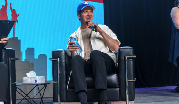 Hayden is wearing a beige shirt underneath a white button up blouse with navy pants. He is also wearing a Toronto Maple Leafs baseball cap. He is holding a water bottle and speaking into a microphone onstage at CALGARY EXPO.