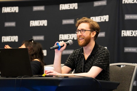 The image captures Justin Briner with glasses, sporting a beard and a black patterned shirt, actively participating in a panel at FAN EXPO. He is holding a microphone and laughing, showing his enjoyment and engagement with the audience or fellow panelists. Behind him, the FAN EXPO logo is prominently displayed across the backdrop, emphasizing the event’s branding. This scene highlights the interactive and vibrant atmosphere typical of such conventions, where panel discussions provide a platform for lively exchanges and community engagement.