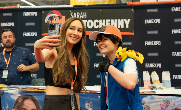 The image captures a cheerful selfie moment at a FAN EXPO event. Sarah Natochenny and a fan are posing in front of a booth, one of whom is dressed in a cosplay outfit resembling a popular anime character, complete with a baseball cap and a Pikachu plush on her shoulder. She's giving a thumbs up, radiating excitement. Sarah, holding the camera for the selfie, has long brown hair and a casual black crop top, smiling broadly. In the background, a man seated at the booth watches the interaction.