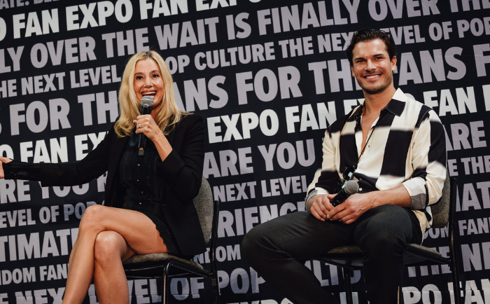Mira Sorvino speaks into a microphone with an animated expression while sitting next to a smiling Gleb Savchenko with dark hair, wearing a black and white striped shirt. Both are seated in front of a backdrop with fan expo text, indicating an event or panel setting. The woman is dressed in a black outfit while the man sports a casual look.