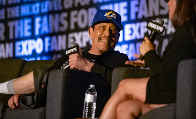 The image shows a lively interaction at a FAN EXPO panel discussion. Danny Trejo, wearing a cap, is laughing expressively, possibly in response to a question or comment. A woman, likely the moderator or interviewer, is holding a microphone and appears to be asking a question or engaging in conversation with him. The man's animated expression suggests a humorous or light-hearted moment. They are both seated in an event setting with a backdrop that reads "FAN EXPO," indicative of a fan convention or cultural event. The focus of the image is on their interaction, capturing a moment of genuine amusement and engagement.