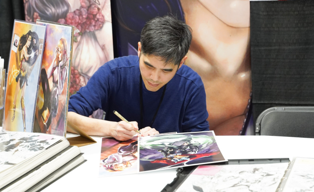 A comic artist works on a new piece at his table. He is signing new coloured art.