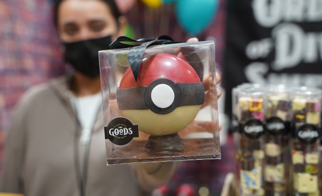 Vendor poses with a chocolate Pokeball in a clear plastic box. The sticker on the box says The GOoDS. More tubes filled with chocolate are in the background.