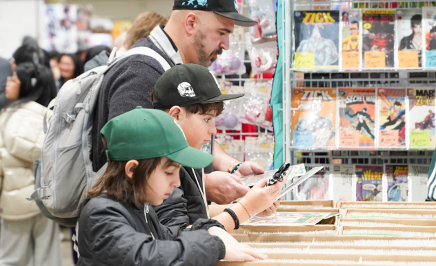 Three people flip through comics at a comic vendor. The comics are in large white boxes.