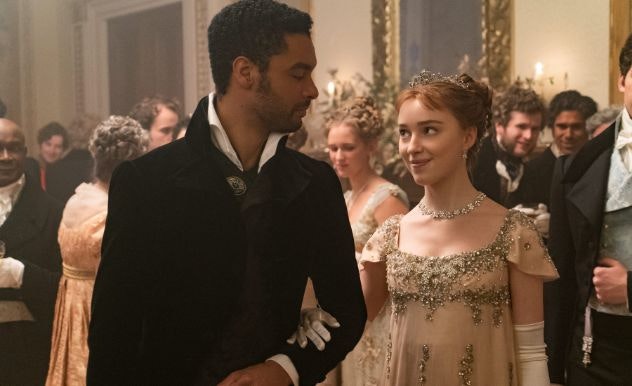 A man in a black tuxedo stands as a woman in a beige dress takes his arm and smiles at him