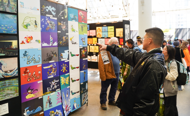 A fan points as he shops an art vendor. They stand in front of the wall of hand painted art, selecting a piece.