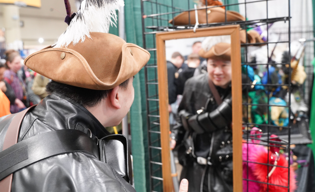 A fan shops for hats at a hat vendor, trying one on and checking out how it looks in the mirror. You see their back to the camera, and their face in the mirror