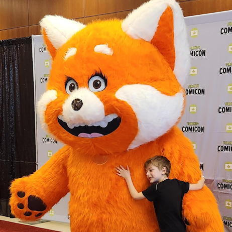 A young boy hugs a cosplayer dressed up as Mei Mei from Turning Red. The Red Panda costume is twice the size of the young boy.