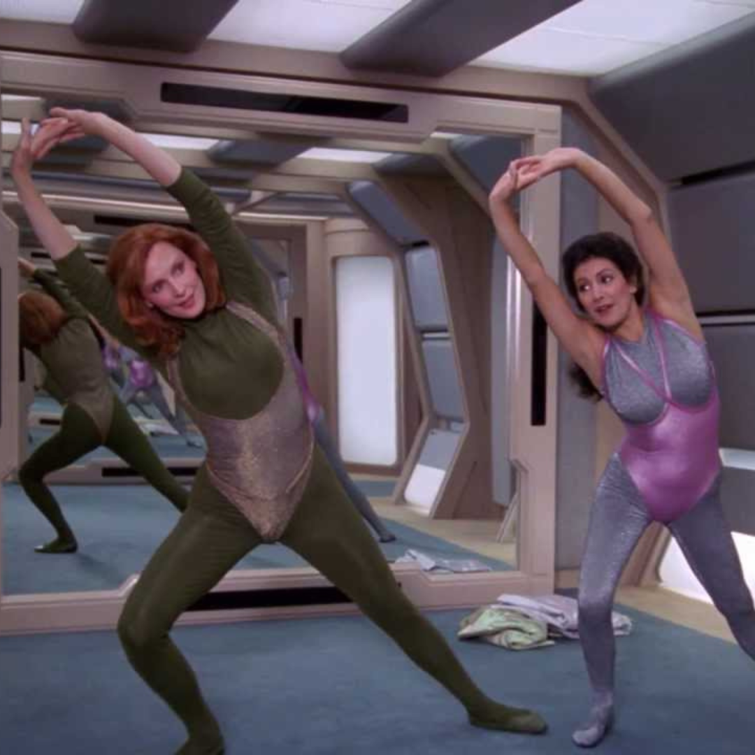 Gates McFadden as Beverly Crusher in a green suit with a silver leotard. Next to her is another woman in a silver suit and a pink leotard. They are stretching in a futuristic dance studios.