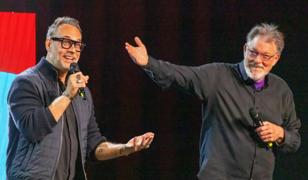 Todd Stashwick and Jonathan Frakes stand on stage holding microphones. Jonathan is pointing at Todd while Todd speaks into a microphone.