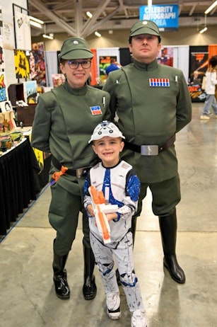 The image shows a family dressed in themed costumes at a fan convention. The two adults are wearing matching green military-style uniforms, complete with hats and medals, reminiscent of characters from a classic film or series. Between them stands a young boy, smiling broadly, dressed in a white and blue costume that appears to be inspired by a popular science fiction character. He holds a toy blaster, adding to the playful and imaginative aspect of their outfits. This family portrait exemplifies the fun and shared enthusiasm that such events foster among attendees of all ages.