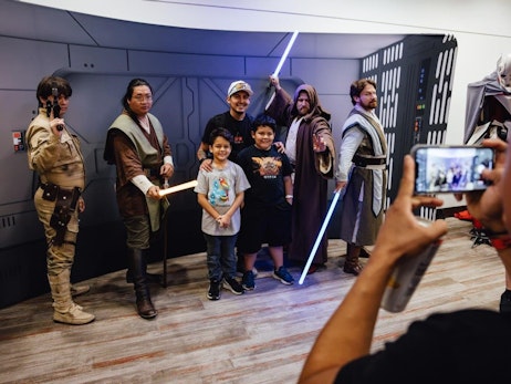 A group of individuals posing in front of a spaceship-like backdrop. On the left, there's a person dressed as a "Star Wars" character in tan military-style attire holding a communication device. Beside them stands another individual portraying a Jedi, with dark hair tied back, wearing brown robes, and holding a lit-up yellow lightsaber. Next to the Jedi, two smiling children stand, with one of them wearing a shirt with a colorful design. On the right, two more individuals dressed as Jedi pose with their lightsabers, one blue and one not illuminated. In the foreground, a person's hands are seen holding a smartphone, capturing the moment on camera. The overall mood is cheerful and celebratory, capturing the essence of a fan event.