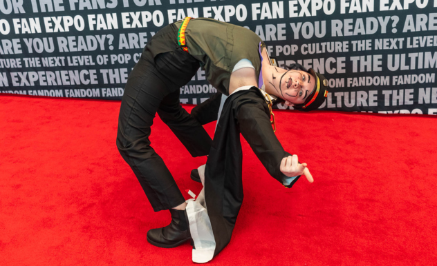 The image features an individual in a playful and dynamic pose on a red carpet, bending backwards with one hand supporting their weight and the other giving a thumbs-up. They are dressed in a costume that includes black pants, a long black coat with gray details, and a hat with goggles, suggesting a character that may be associated with aviation or steampunk themes. The person's attire is completed with a multicolored striped scarf and a joyful expression, adding to the whimsical nature of the pose.