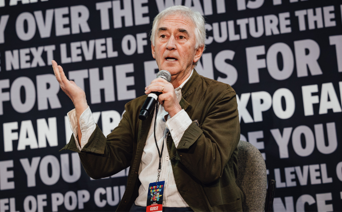 Denis Lawson with silver hair speaks into a microphone while gesturing with his hand. He wears a white shirt underneath a dark green jacket and has a badge labeled "GUEST" hanging from his neck. He sits in front of a backdrop with repeated fan expo text, indicating he's at a panel or event setting.