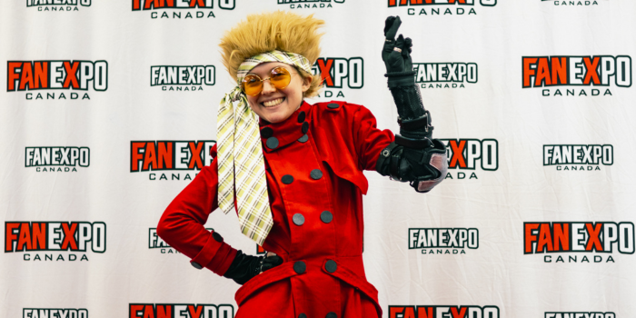 Cosplayer poses with hand on hip and one hand up, bent at the elbow. They are dressed as Vash the Stampede with a tie tied around their head.