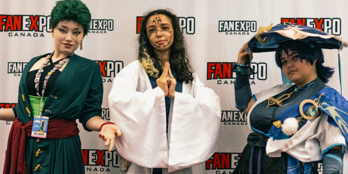 Three fans pose together as characters from various animes. The first, holds their hands out to either sides like a shrug, the second has their hands folded, index, middle, and ring fingers pointed upward and touching, and the third tips their hat away from the camera.