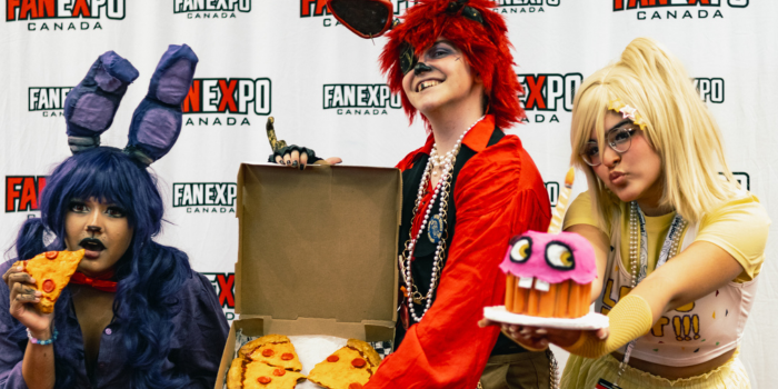 Three fans dressed as personified Five Nights at Freddy's characters. Left to right: Bonny (eating a slice of pizza), Foxy (holding a box with pizza slices), and Chica, holding the cupcake.
