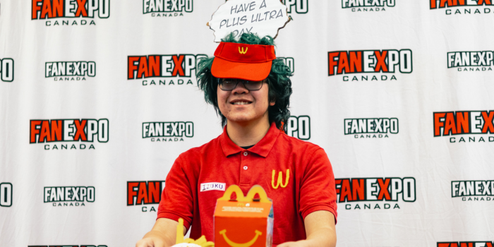 Dressed as a WcDonald's employee, a fan holds out a take out tray with a faux meal on it. Above their head is a speech bubble that reads "have a Plus Ultra day!"