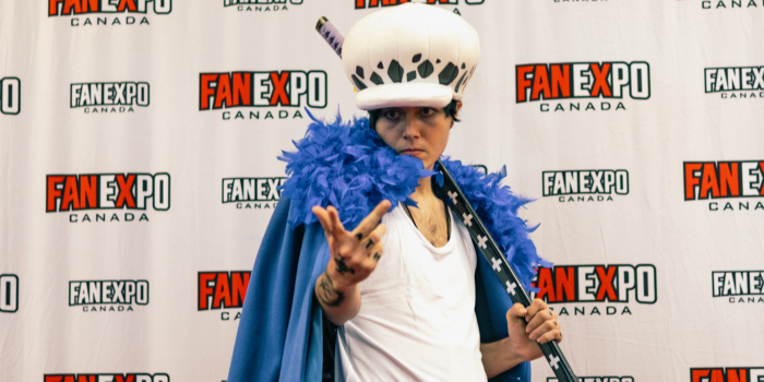 A fan stands with their arm extended toward the camera, dressed as Trafalgar D. Water Law from One Piece. They are holding their staff in their opposite hand, leaning it behind their head. 