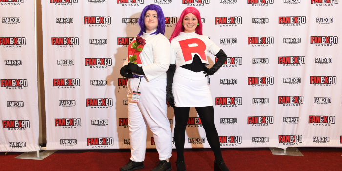A couple poses, angled back to back - they are dressed as Jessie and James from Pokemon. James is holding a red rose. 