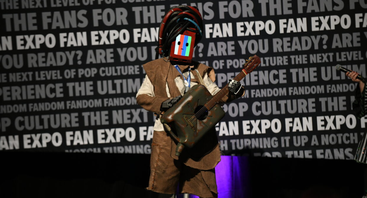 Morusqe from Stray. The head is a functional screen with tangled wile hair. The cosplayer holds a guitar built from an old gas can.
