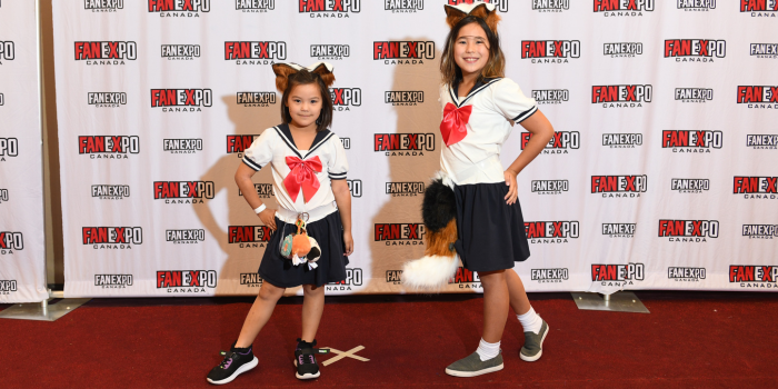 Two young sisters dressed in identical outfits are posed with their hands on their hips, sporting calico coloured fox ears and tails.